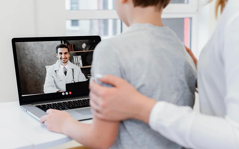 Consulta médica por videollamada en una laptop mientras una madre acompaña a su hijo